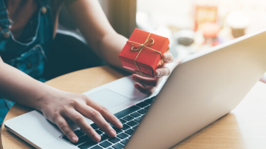 Woman's hand entering information into a laptop while holding a red gift box in her hand.
