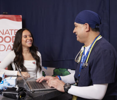 CBC doctor answering questions for a blood donor with a sign reading Donate Blood Today in the background.