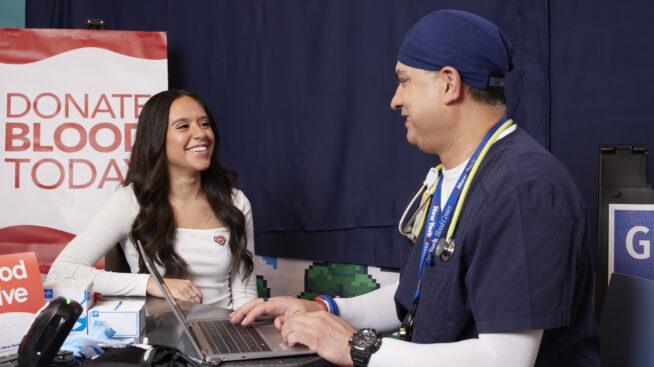 CBC doctor answering questions for a blood donor with a sign reading Donate Blood Today in the background.