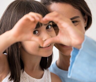 Smiling ittle girl and her mother peering through the girls heart shaped hands.